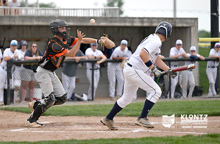 Baseball catcher in black and orange gear lunges for a high throw as a batter in white runs toward home plate with spectators in the background.