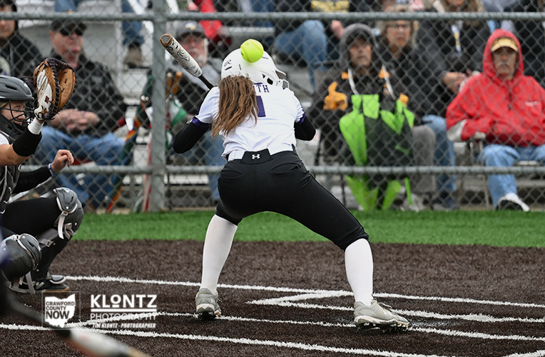 Female softball batter in white uniform (#1) at home plate, ready to swing as the neon-green ball approaches; catcher and crowd in the background.