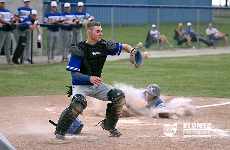 Baseball catcher in blue, black gear, blocks home as a baserunner slides in and dust rises around the plate