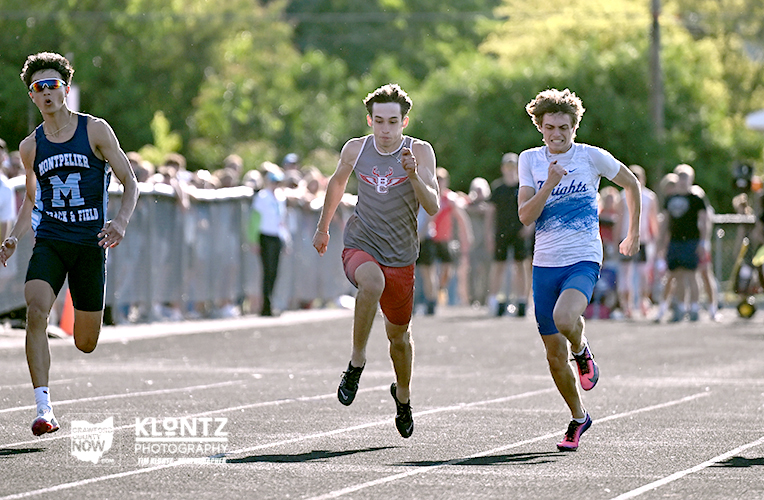 Three male sprinters racing toward the finish line on a track with a cheering crowd behind them.