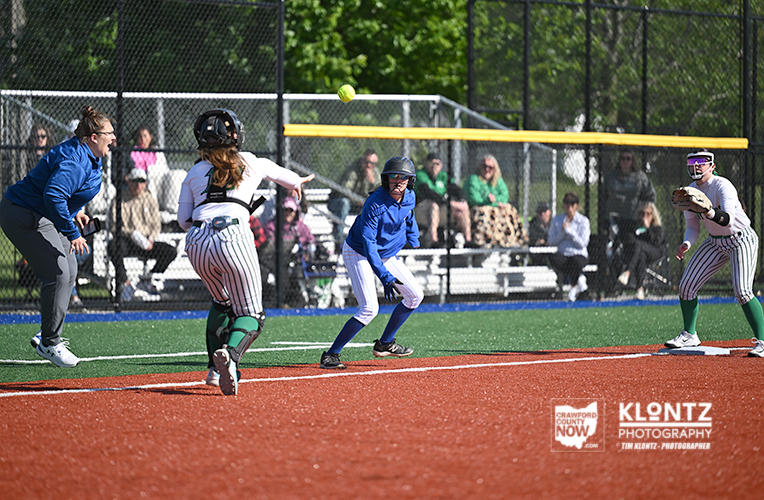 Youth softball game: blue-uniform pitcher delivers a pitch toward home as catcher and baseman watch, with spectators behind the fence.