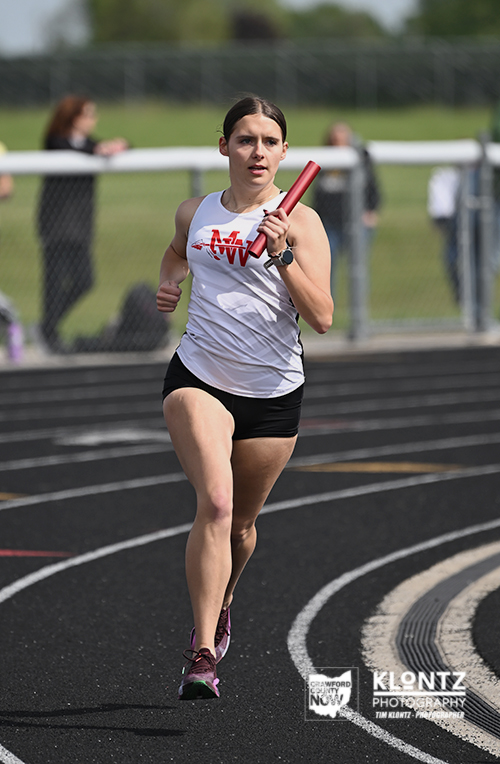 Female sprinter on a relay track sprinting with a red baton in her right hand under a sunny sky.