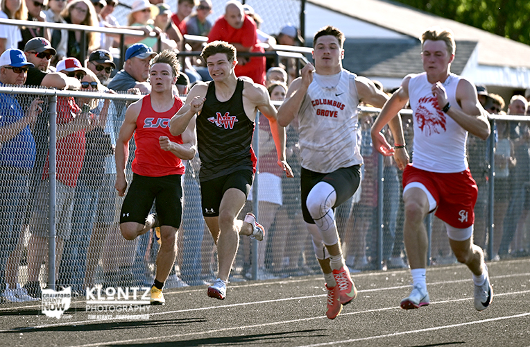 Four male sprinters race toward the finish on an outdoor track, with a cheering crowd behind a chain-link fence.
