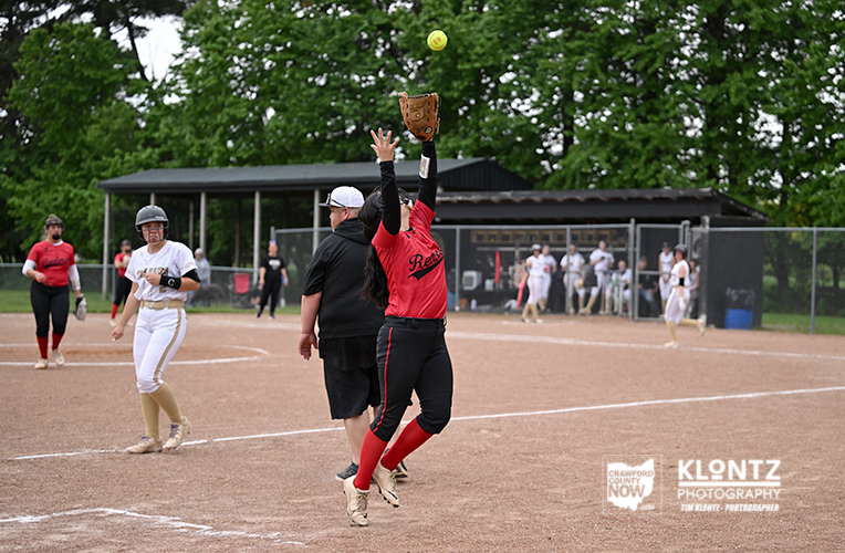 Female softball player in a red uniform jumps to catch a ball with her glove raised; teammates and dugout in the background.