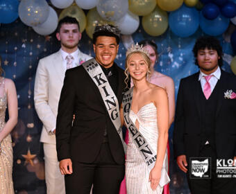 Prom king and queen pose center stage, wearing crowns and sashes, with balloons and classmates in formal attire in the background.