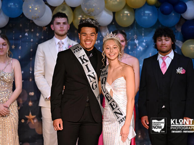 Prom king and queen pose center stage, wearing crowns and sashes, with balloons and classmates in formal attire in the background.