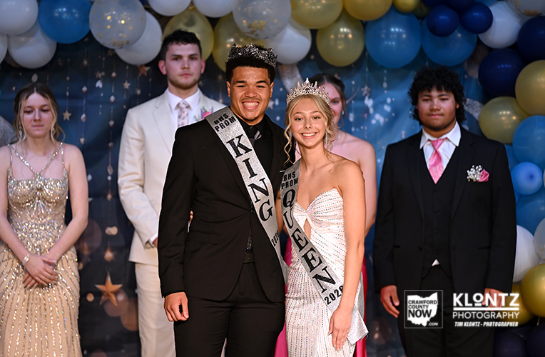 Prom king and queen pose center stage, wearing crowns and sashes, with balloons and classmates in formal attire in the background.