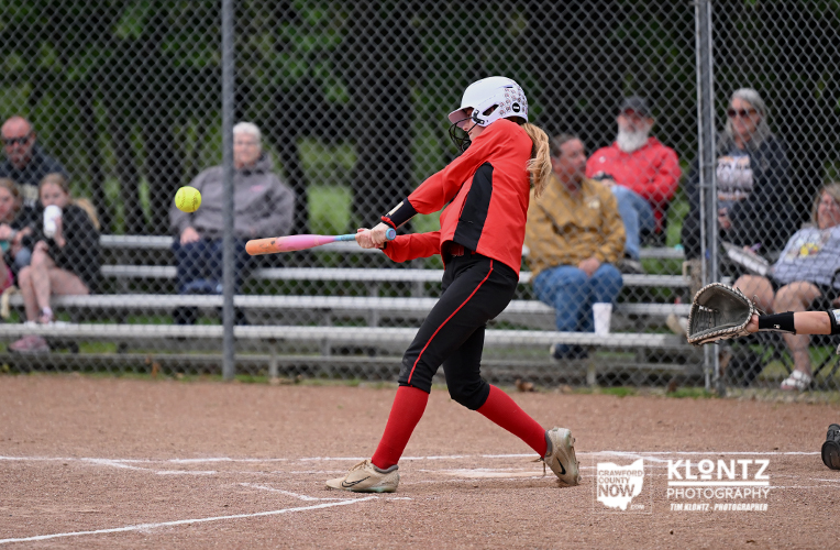 Female softball player in red and black swings a bat at a pitched ball on a dirt field, with spectators behind a chain-link fence.