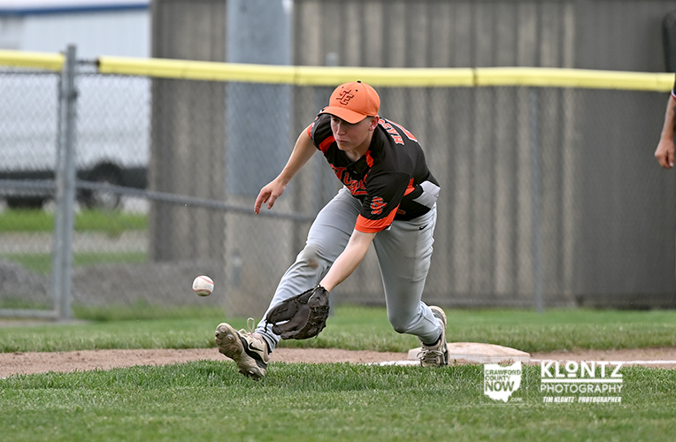 Young baseball player in black-orange uniform dives to scoop a ground ball near first base, glove extended toward the ball with orange cap on.