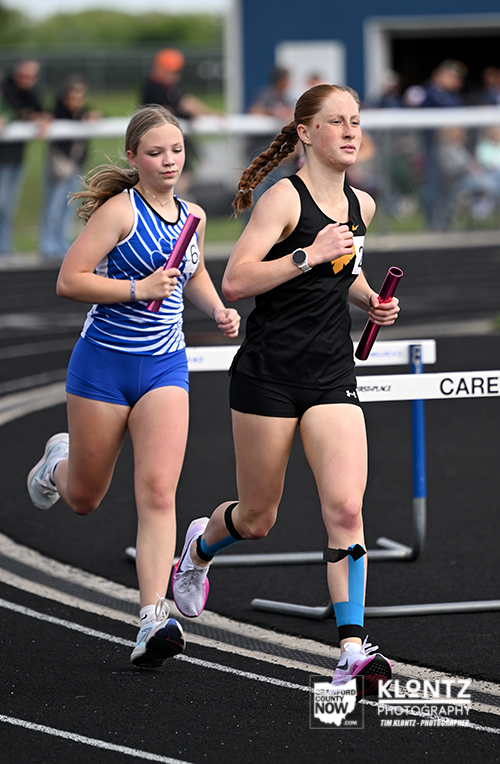 Two young female sprinters with batons run on an outdoor track, one in a blue striped uniform and the other in black, passing a hurdle as spectators watch in the background.