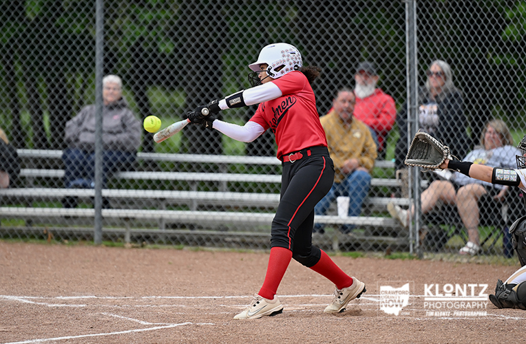 Female softball batter in a red uniform swings at a pitched ball near home plate, with catcher nearby and spectators behind a chain-link fence.