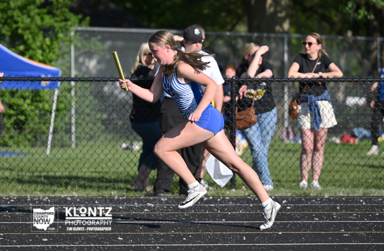 Female relay runner sprinting on a track with a baton, teammates and spectators behind a chain-link fence in a sunny meet scene