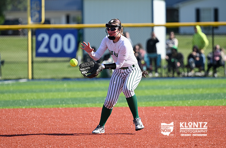 Female softball fielder in white jersey and striped pants reaches for a low ball with glove on a red infield, spectators in the background.