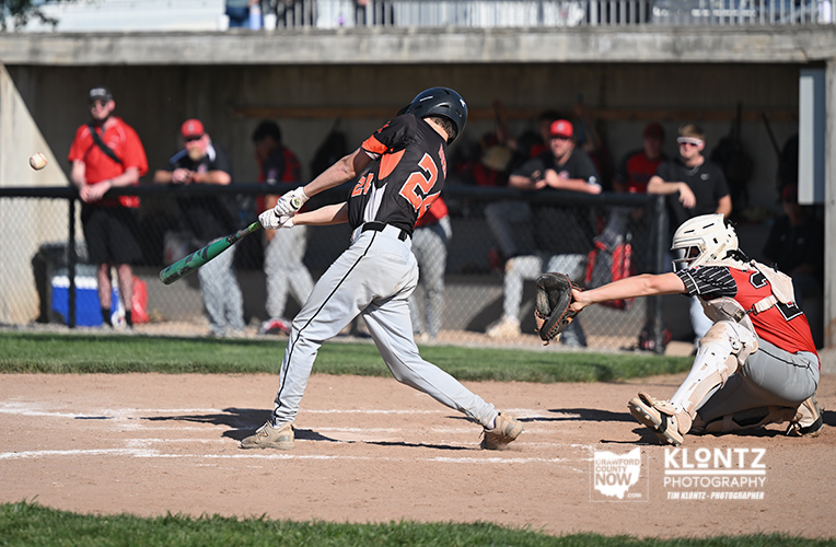 Baseball action: batter in black and orange swings at a pitch as the catcher in red and white lunges to stop the ball near home plate.