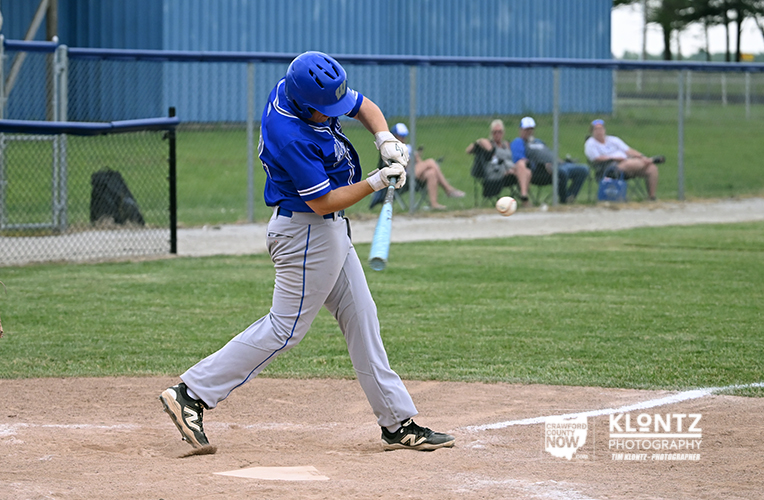 Baseball batter in blue uniform swings at a pitch near home plate with onlookers in the background.