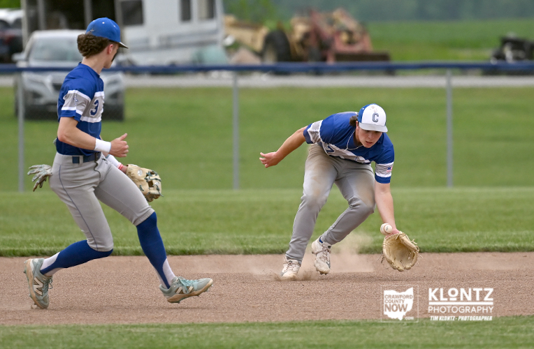 Two baseball players in blue and gray uniforms chase a ground ball on the infield; one slides while the other approaches with a glove ready.