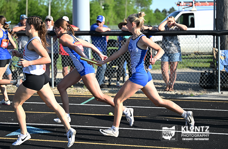 Female athletes in blue uniforms relay race on track; one hands off baton to teammate as others run along the lane edge on a sunny day.