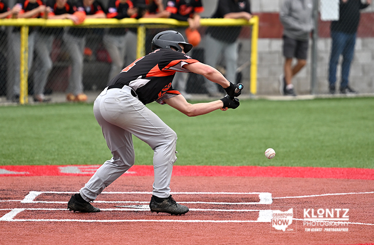 Baseball player in black-and-orange uniform crouches at home plate, swinging at an incoming pitch as the ball approaches.