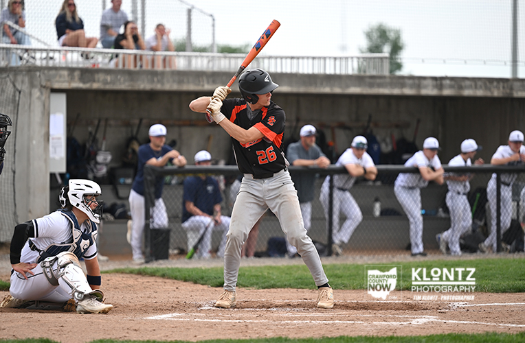 Young baseball batter in a black-and-red jersey swings his orange bat at a pitch, with a catcher in white gear squatting behind home plate.