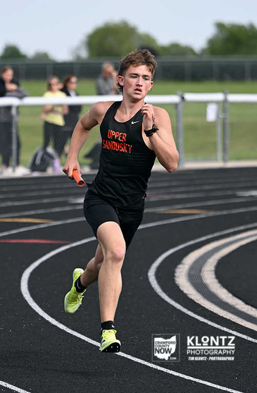 Young male track sprinter wearing a black Upper Sandusky uniform runs on a curved outdoor track, holding an orange baton as spectators watch from behind a fence.