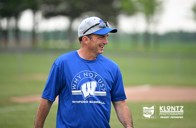Man in a blue Wynford Baseball shirt smiles while walking on a baseball field outdoors.