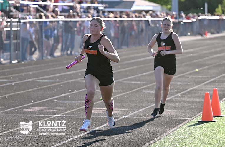 Two female sprinters race on a track; the front runner in black holds a pink baton as spectators watch from behind a fence.