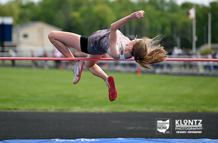 Female high jumper in mid-air clearing the bar during a track and field event on a green field.
