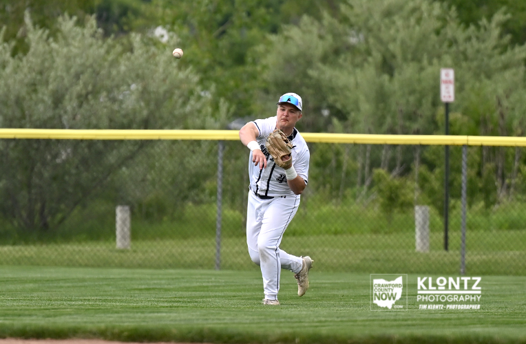 Baseball pitcher in a white uniform throws a pitch on a grassy field with a yellow outfield fence behind him, trees in the background as the ball flies away.