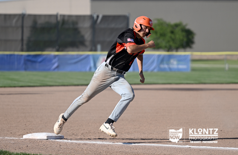 Baseball baserunner sliding toward first base on a dirt infield, wearing an orange helmet and black/orange uniform with a white cleat close to the bag.