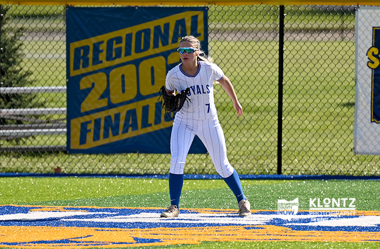 Female softball player in white and blue uniform standing in the infield, glove ready, with a Regional Final banner in the background on a chain-link fence.