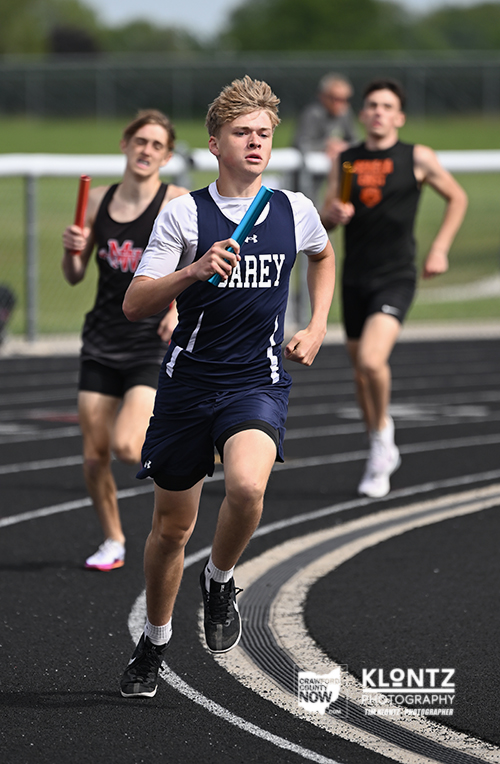Young male sprinter in a navy blue relay uniform holding a turquoise baton leads a race on an outdoor track, with two pursuing runners behind him.