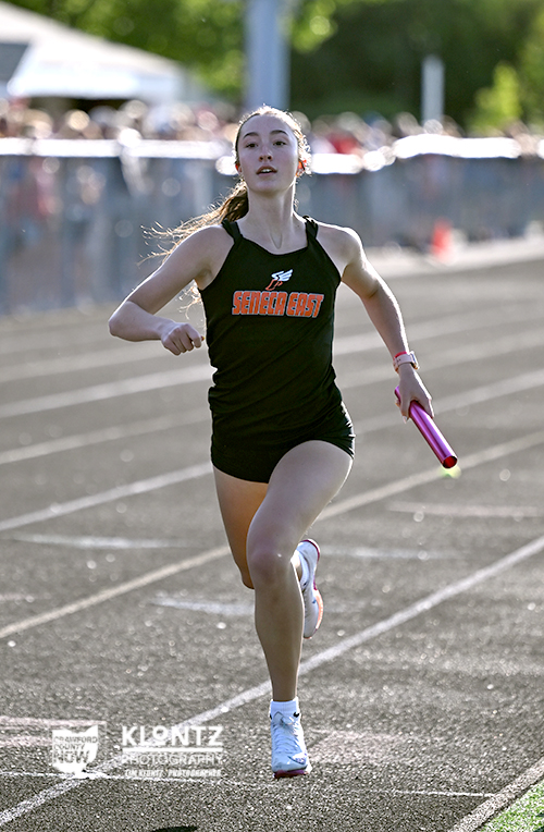 Female relay runner in a black Seneca East uniform sprinting on a track with a pink baton.