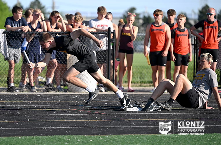 Male sprinter in black uniform lunges to pass a baton to a teammate on a track during a relay; crowd watches behind a chain-link fence.