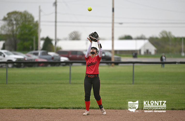 Young softball player in red uniform catches a pop-up on a dirt infield, glove raised, ball in the air above her.