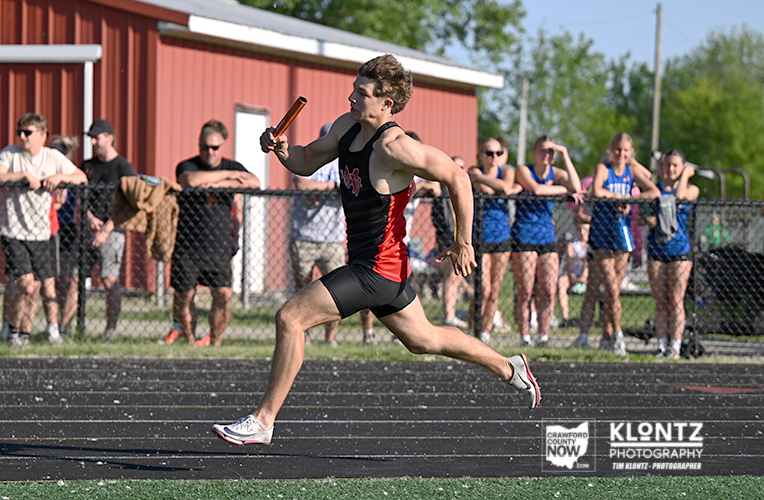 Relay race: male sprinter in black and red uniform runs with baton on a track, spectators behind a chain-link fence behind him.