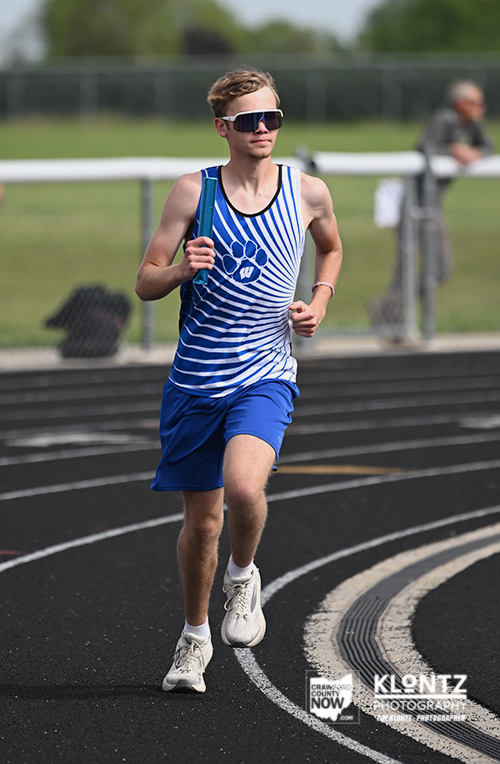 Young male athlete running on an outdoor track in a blue striped singlet and blue shorts, wearing sunglasses and carrying a water bottle strap over his shoulder.