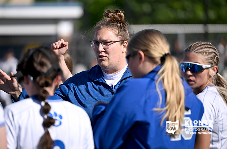 Coach in a blue jacket speaks to a girls' sports team during practice on a sunny field with a chain-link fence in the background.