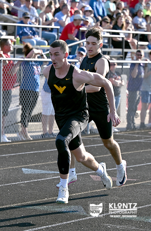 Two male sprinters race on a track, front runner wearing a knee brace and black singlet with a yellow logo, crowd in stands behind them