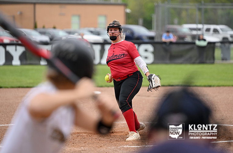 Female softball pitcher in red 'Redmen' jersey throws a pitch on a dirt mound, glove ready, with a bright yellow softball in hand. Behind her are blurred players and a fence with banners on a grassy field.