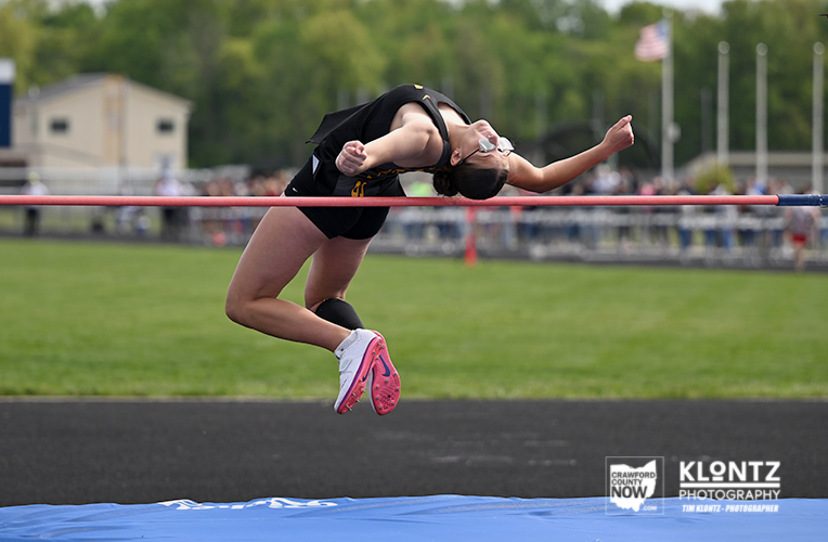 Female high jumper clears the bar mid-air at an outdoor track meet, landing on a blue mat.