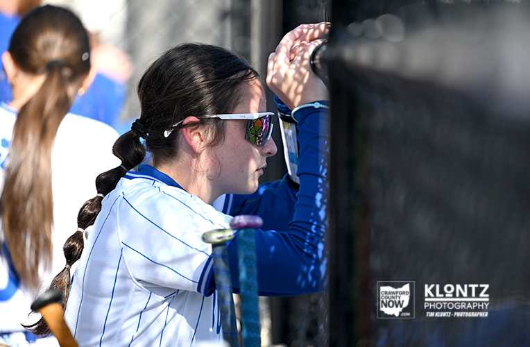 Female athlete in a white-blue jersey with a braided ponytail, wearing sunglasses, adjusting gear beside a team bus.