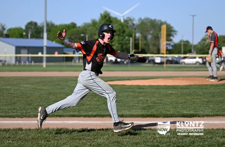 Young baseball player in a black and orange uniform sprints toward first base on a sunny day, wearing a helmet and glove as the field and a teammate/coach stand in the background.