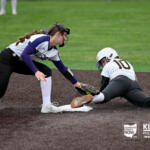 Softball player slides into first base as fielder reaches to tag her at the bag on a dirt infield