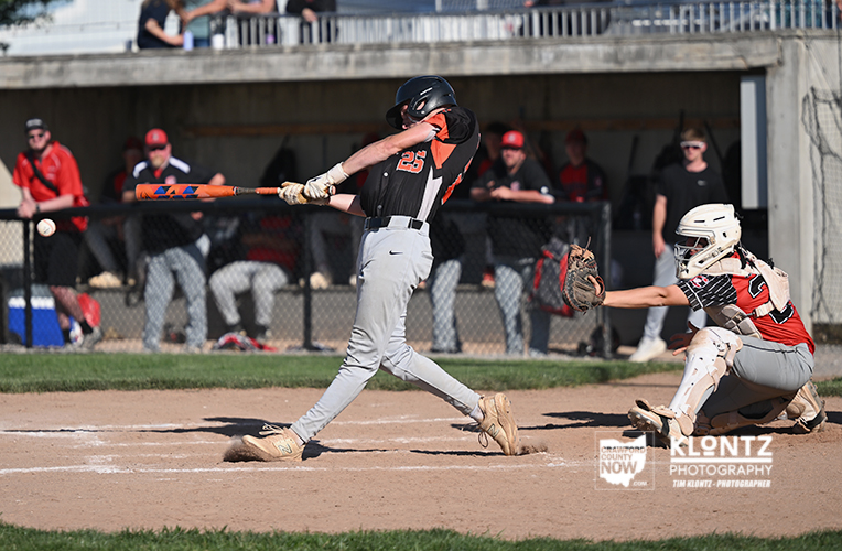 Right-handed baseball batter in black and gray swings at a pitch as the ball nears the bat; catcher in white gear is set behind home plate.