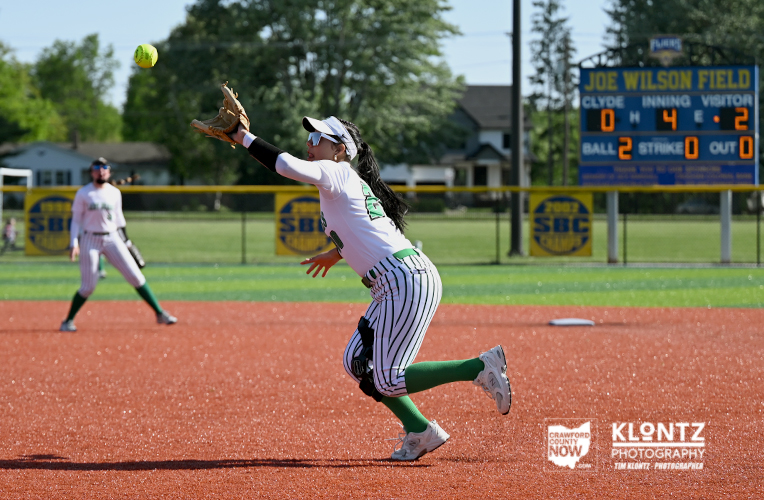 Female softball pitcher in a white pinstripe uniform winds up to throw a ball on a red dirt field, with a scoreboard and another fielder in the background.