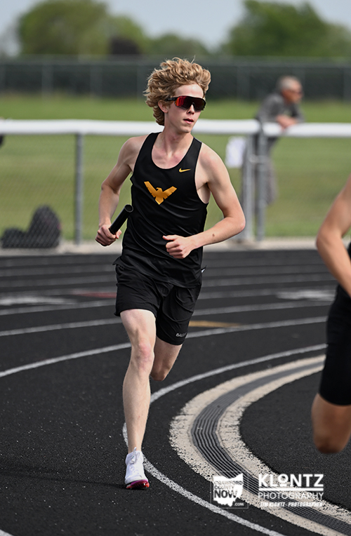 Male sprinter in black athletic gear running on track, holding a baton, wearing red-tinted sunglasses.