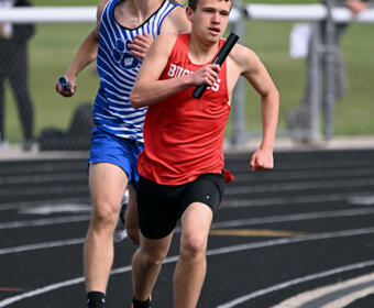 Two boys sprint on a track in a relay race; red-shirt runner leads with a baton, blue-shirt runner close behind.