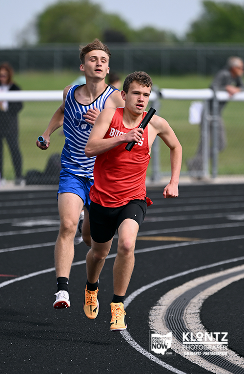 Two boys sprint on a track in a relay race; red-shirt runner leads with a baton, blue-shirt runner close behind.