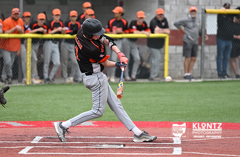 Youth baseball player in a black and orange uniform swings at a pitched ball at home plate as spectators in orange watch from behind a chain-link fence behind the field.