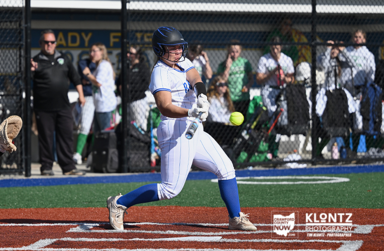 Female softball batter in white pinstripes swings at a pitched ball near home plate, spectators behind a chain-link fence.